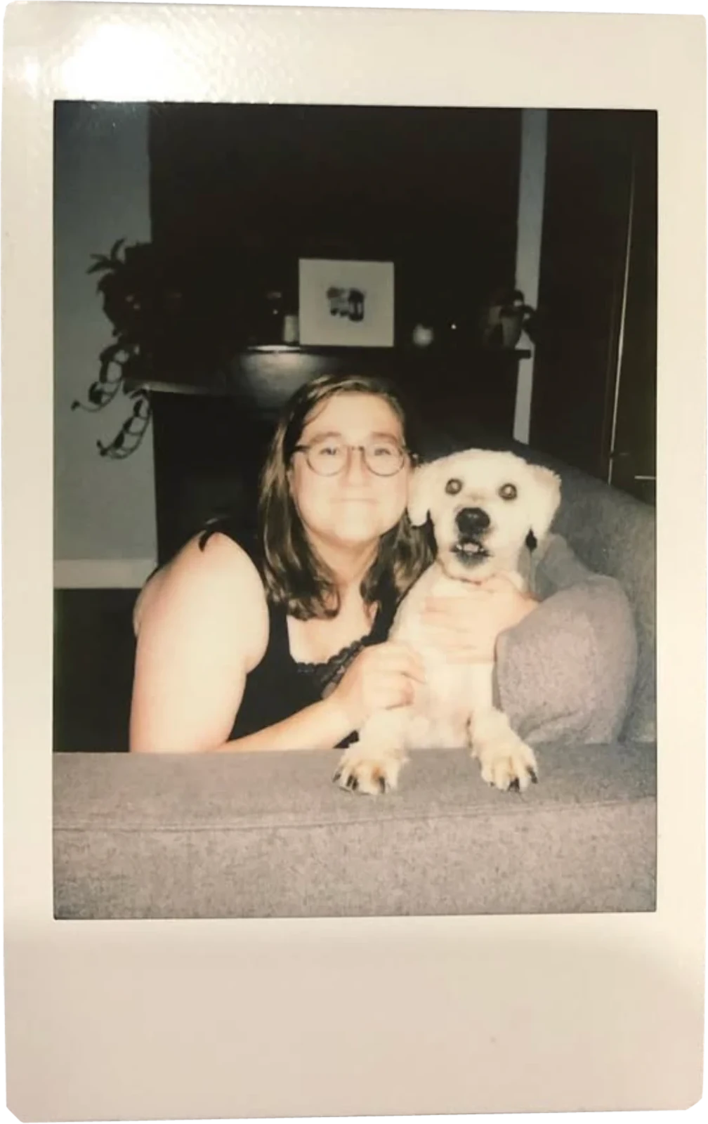 Instant photo of someone in glasses hugging a white dog on a couch