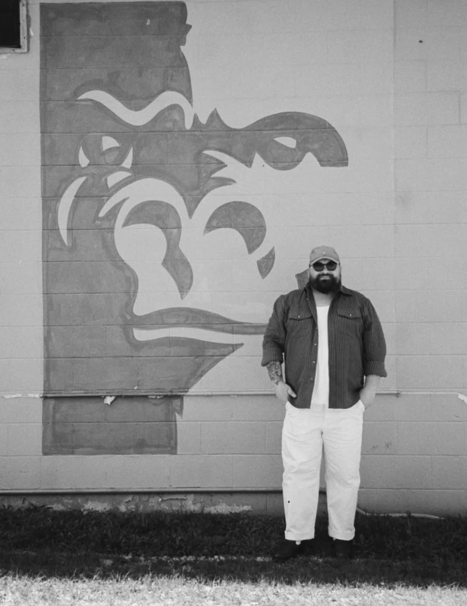A man in a cap and sunglasses beside a painted wall mural, in black and white