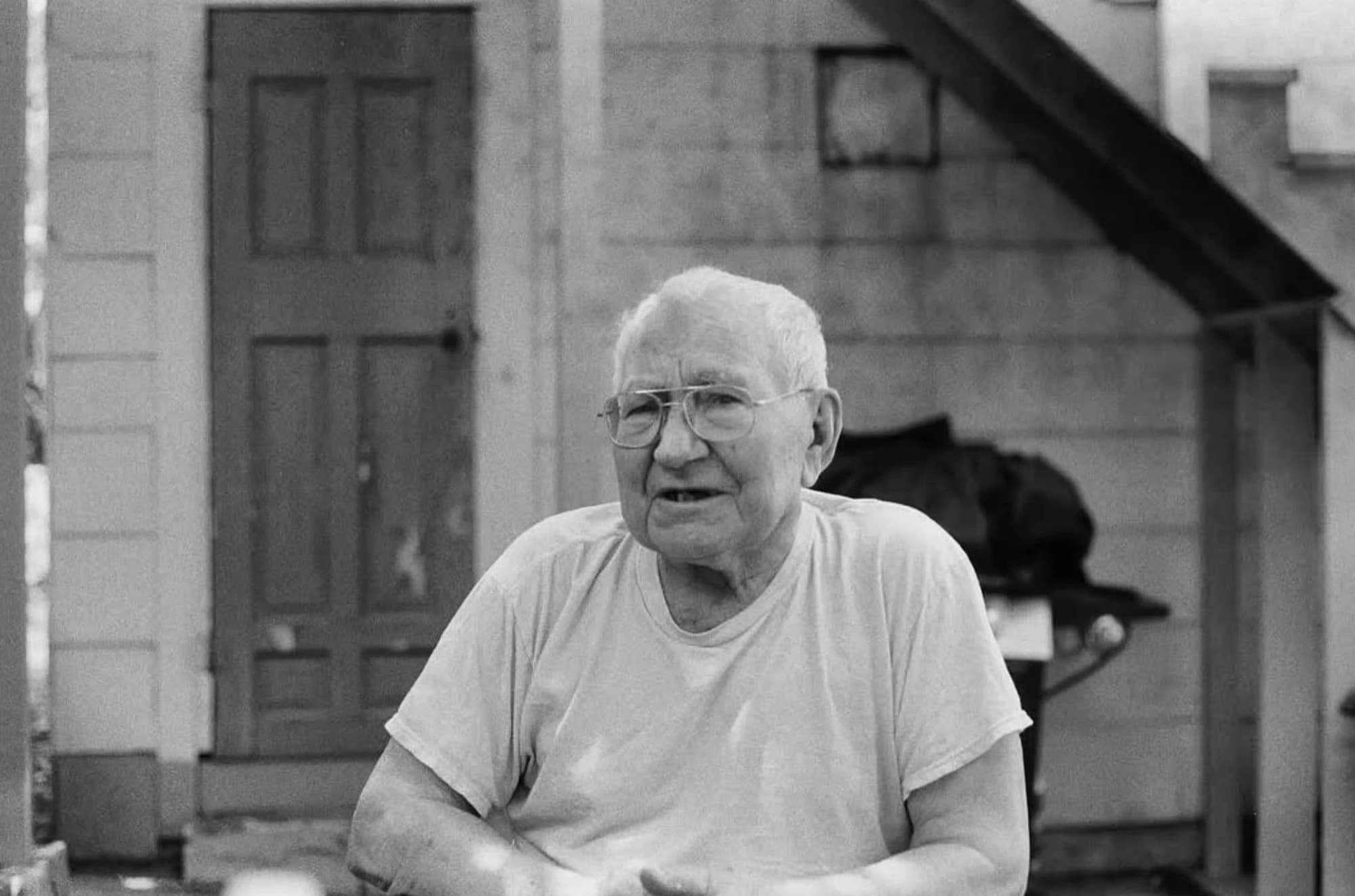 An older man in glasses and a white T-shirt talking outside a house, in black and white