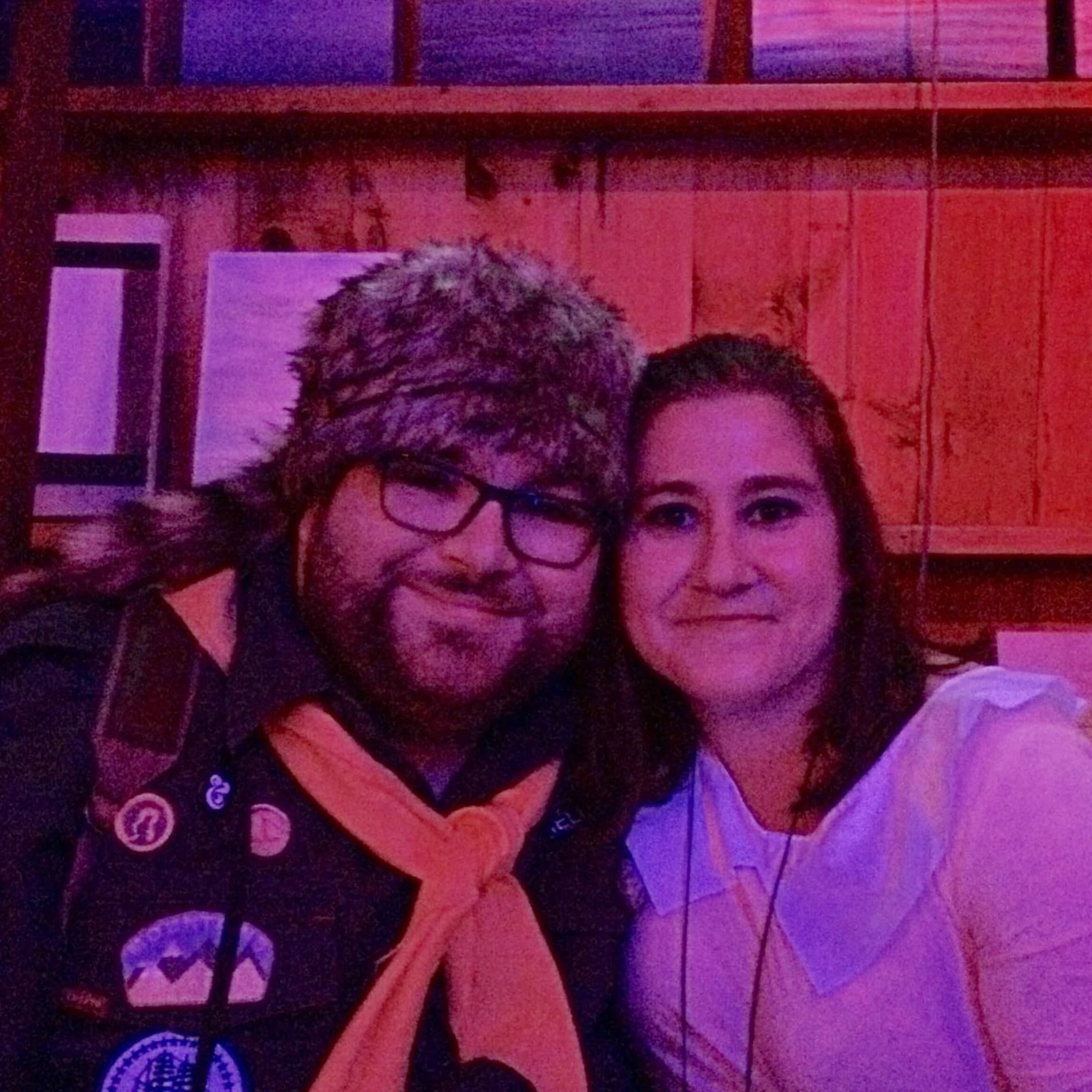 A couple leaning together in red and purple light, one in a fur hat and scout neckerchief