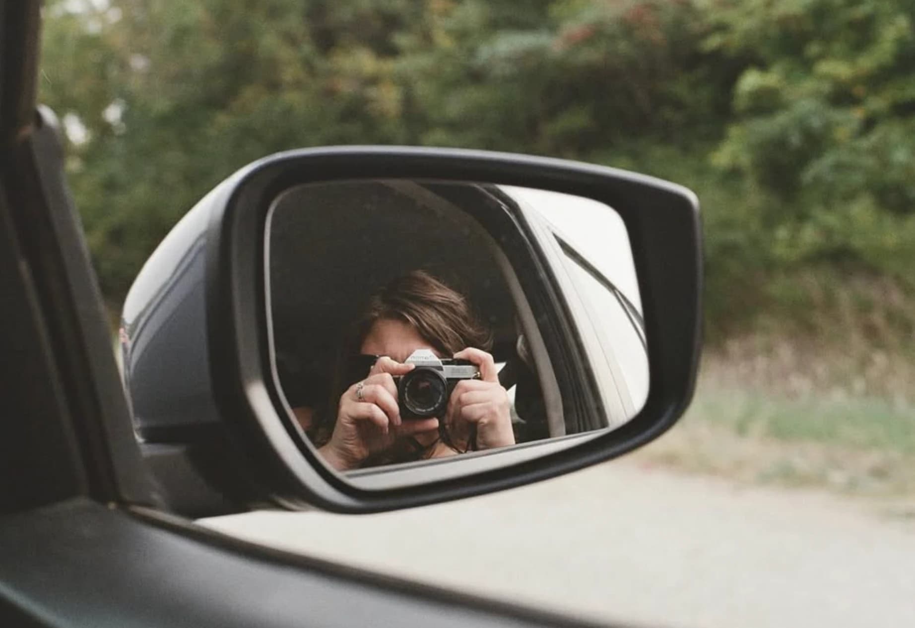 A photographer with a film camera reflected in a car's side mirror